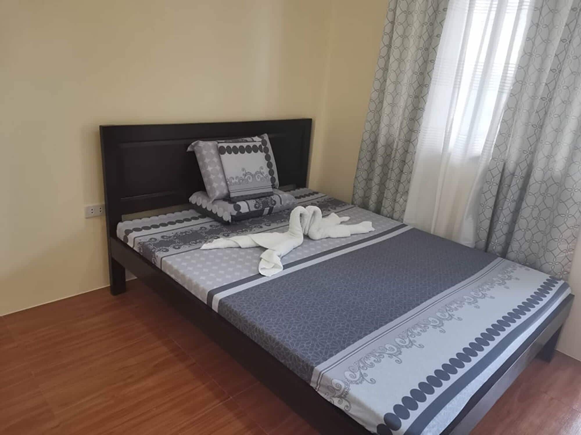 A neatly made bed with patterned bedding and pillows, a dark headboard, and a window with patterned curtains.