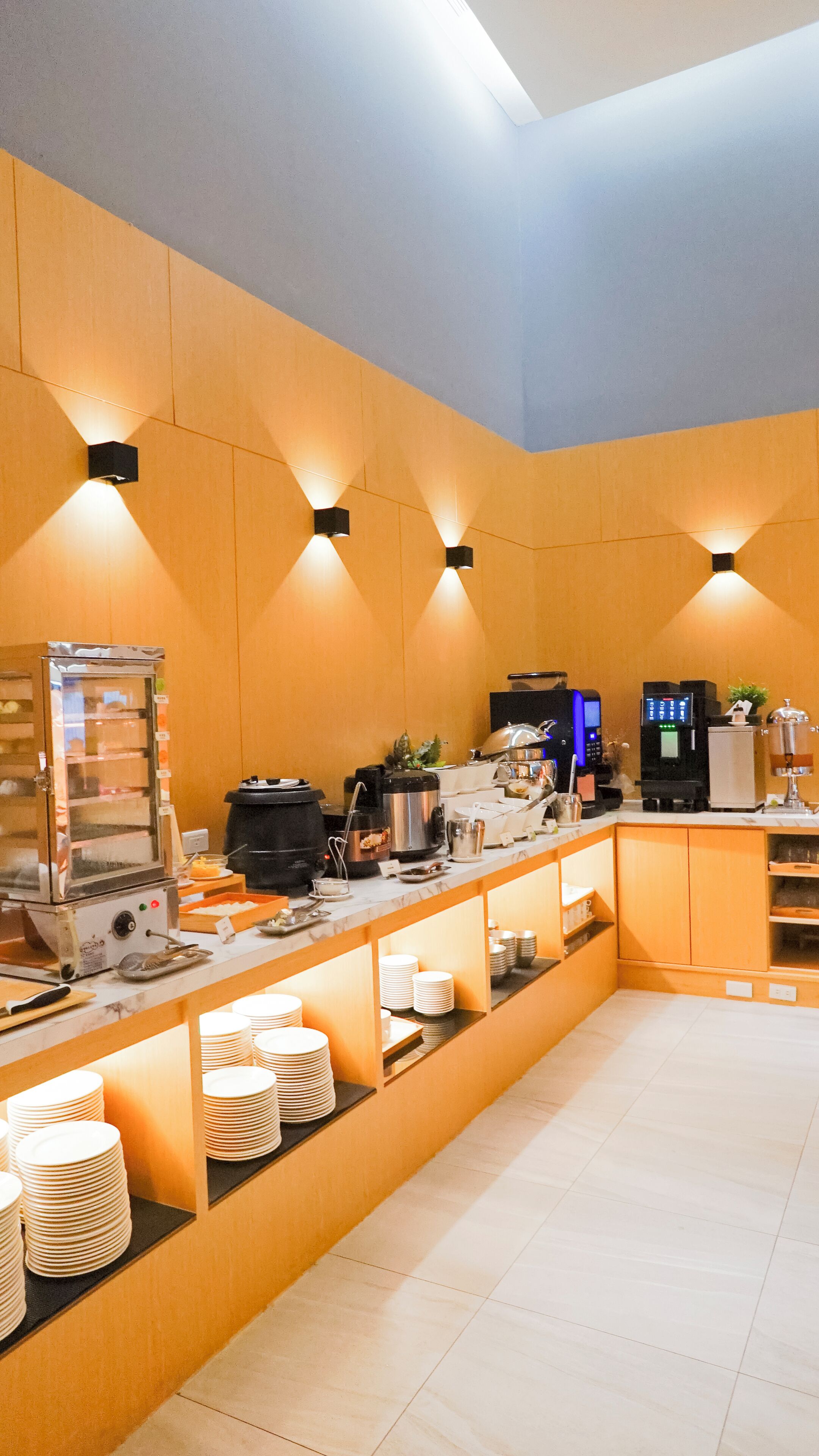 A modern kitchen area with a buffet setup, including a refrigerator, coffee machine, and various food items.