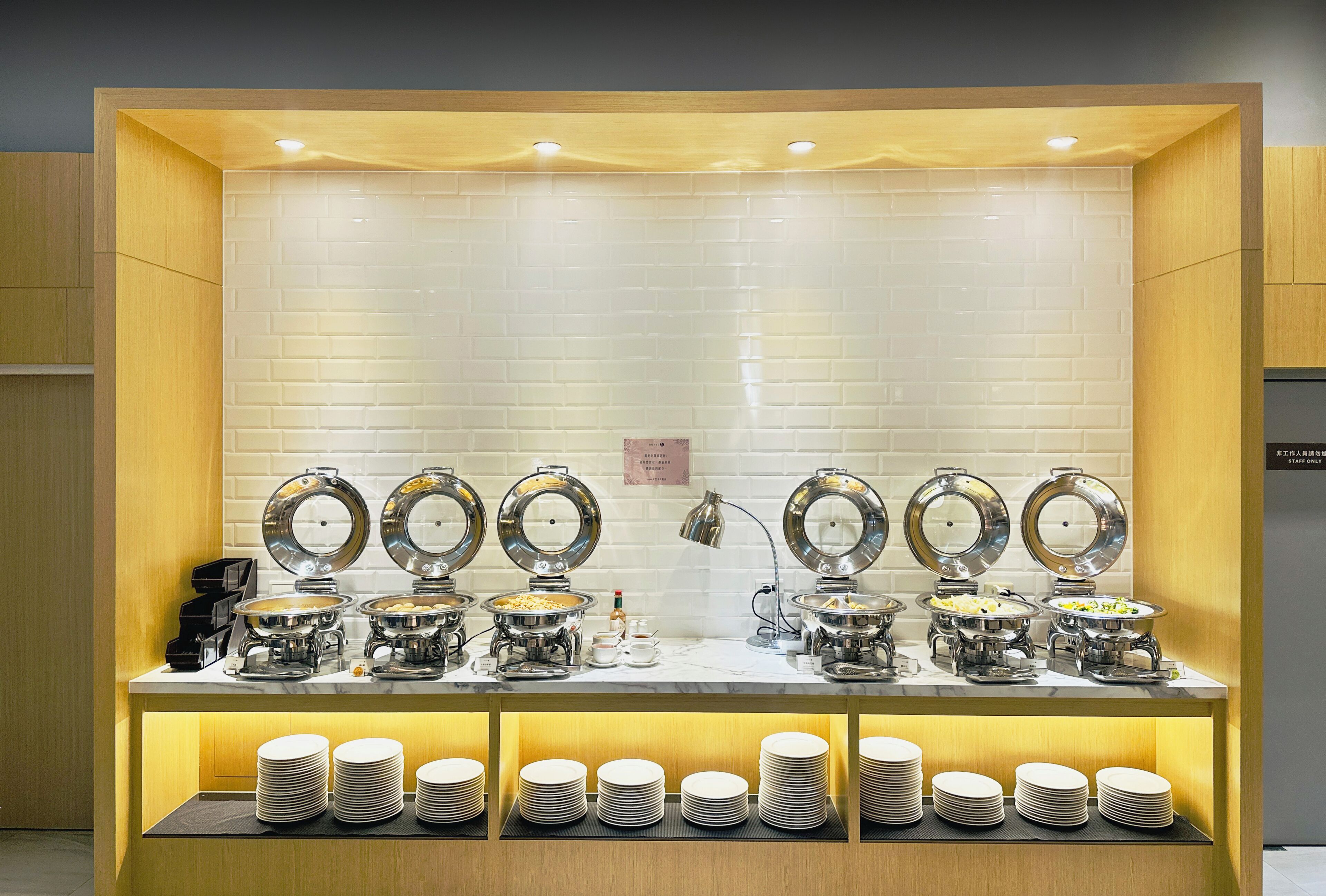 A buffet setup with multiple serving stations, each with a metal bowl and a lid, and a stack of white plates below.