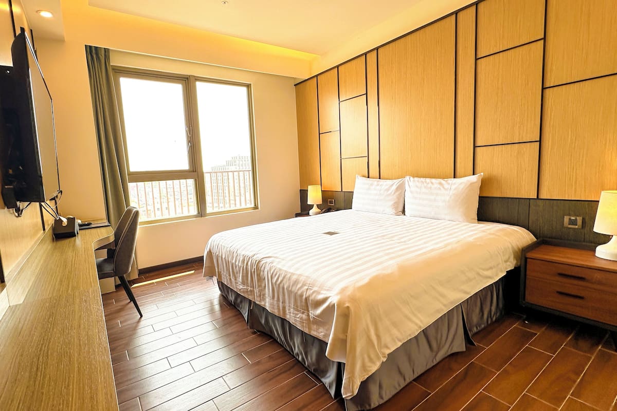 A hotel room with a large bed, a desk, a chair, a TV, a window, and a nightstand with a lamp.