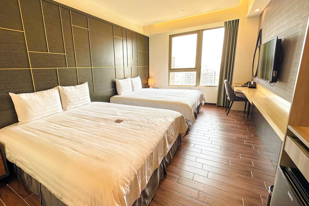 A hotel room with two beds, a TV, a desk, and a window with curtains.