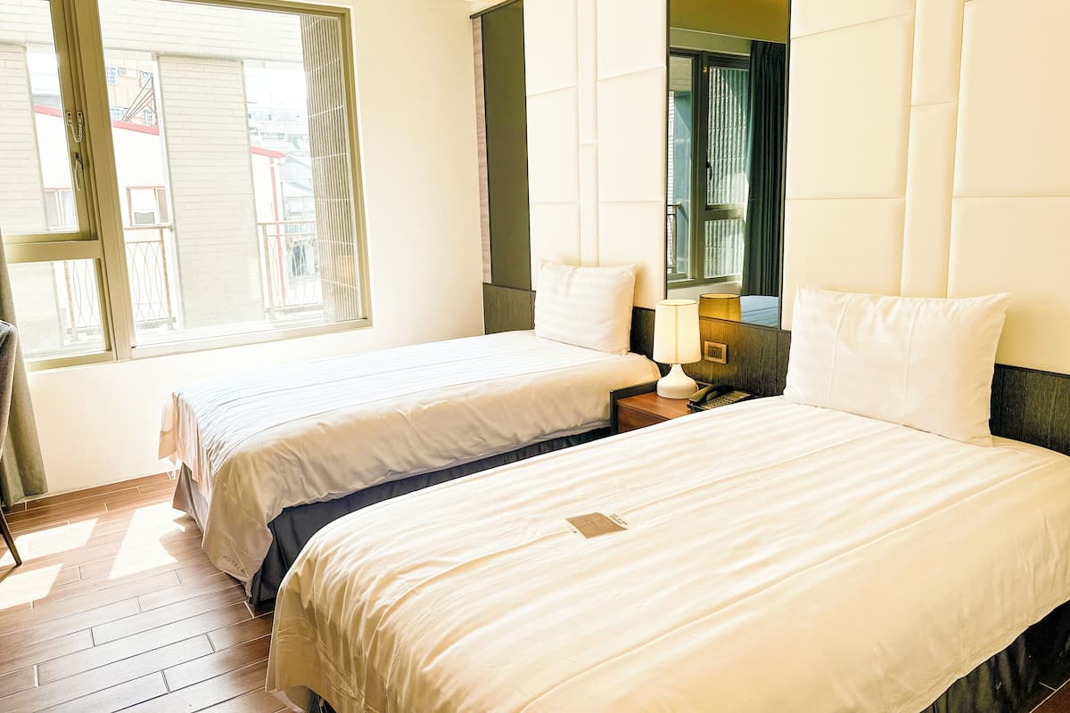 A hotel room with two beds, a large mirror, a bedside table with a lamp, and a window with blinds.