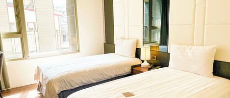 A hotel room with two beds, a large mirror, a bedside table with a lamp, and a window with blinds.