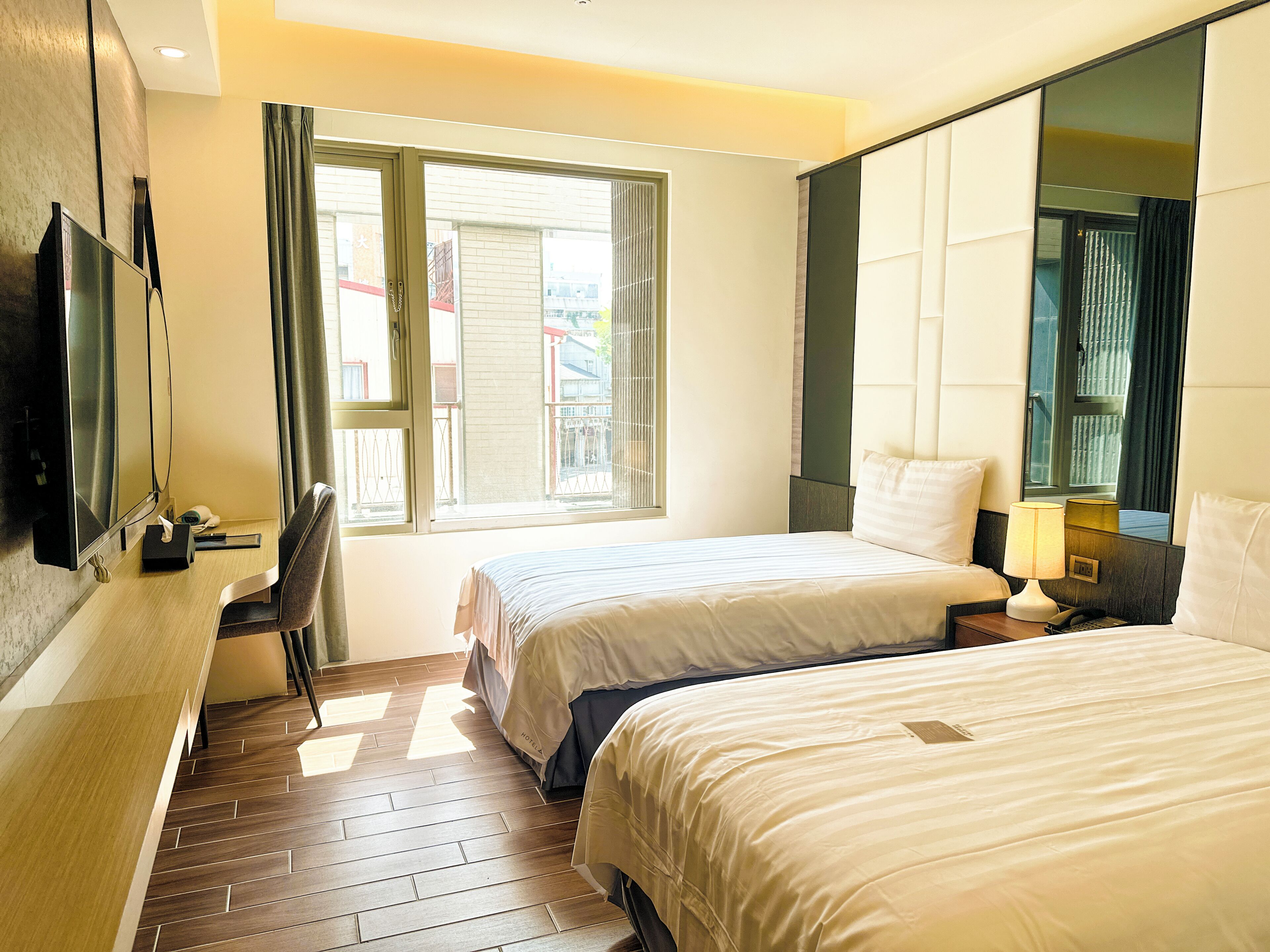 A hotel room with two beds, a TV, a desk, and a window with a view of buildings.