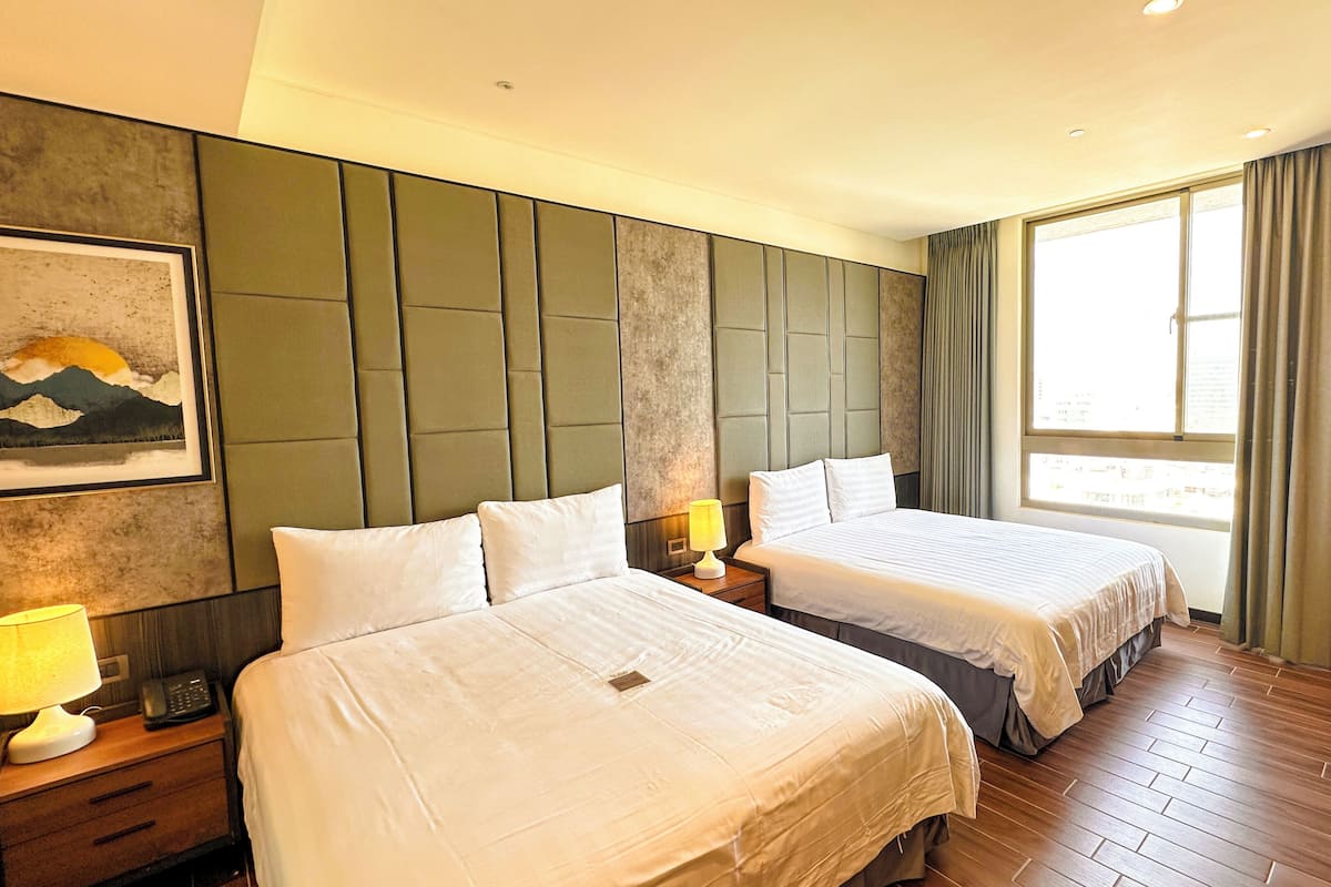 A hotel room with two beds, a desk, a chair, a lamp, a painting, and a window with curtains.