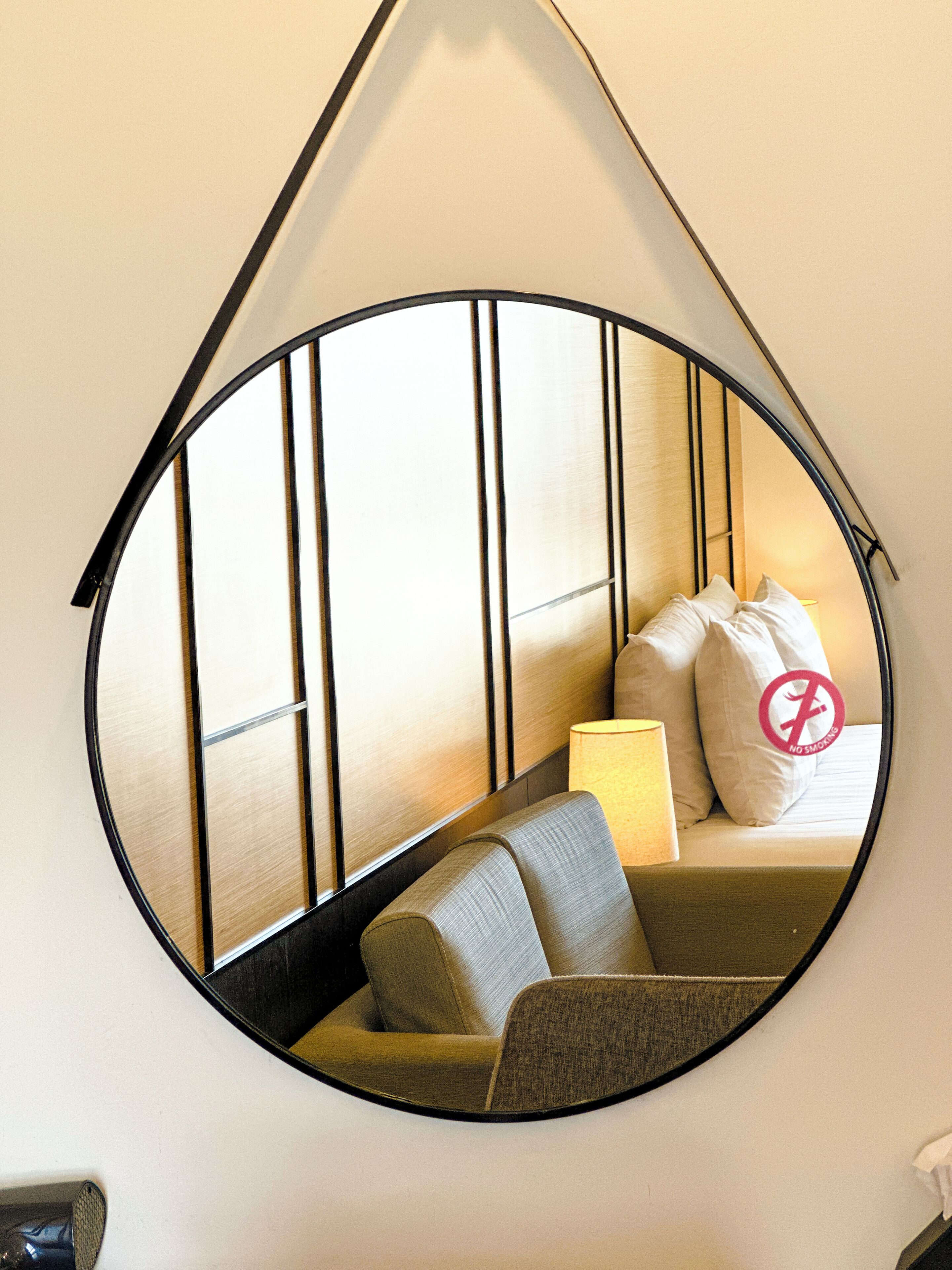 A circular mirror with a black frame, reflecting a room with a sofa, pillows, and a lamp.