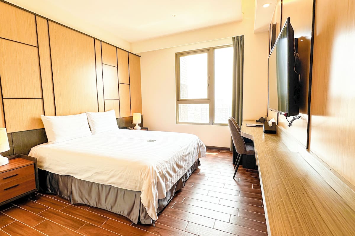 A hotel room with a bed, bedside tables, a desk, a chair, a TV, and a window.