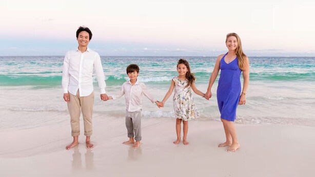 Uma família de quatro pessoas de mãos dadas numa praia, com o oceano ao fundo.
