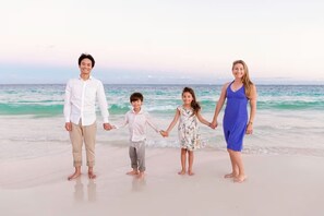 Uma família de quatro pessoas de mãos dadas numa praia, com o oceano ao fundo.
