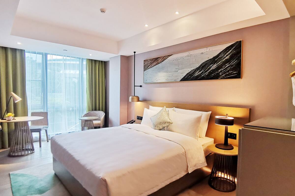 Ein modernes Hotelzimmer mit einem großen Bett, einem Schreibtisch, einem Sessel und Blick ins Freie.