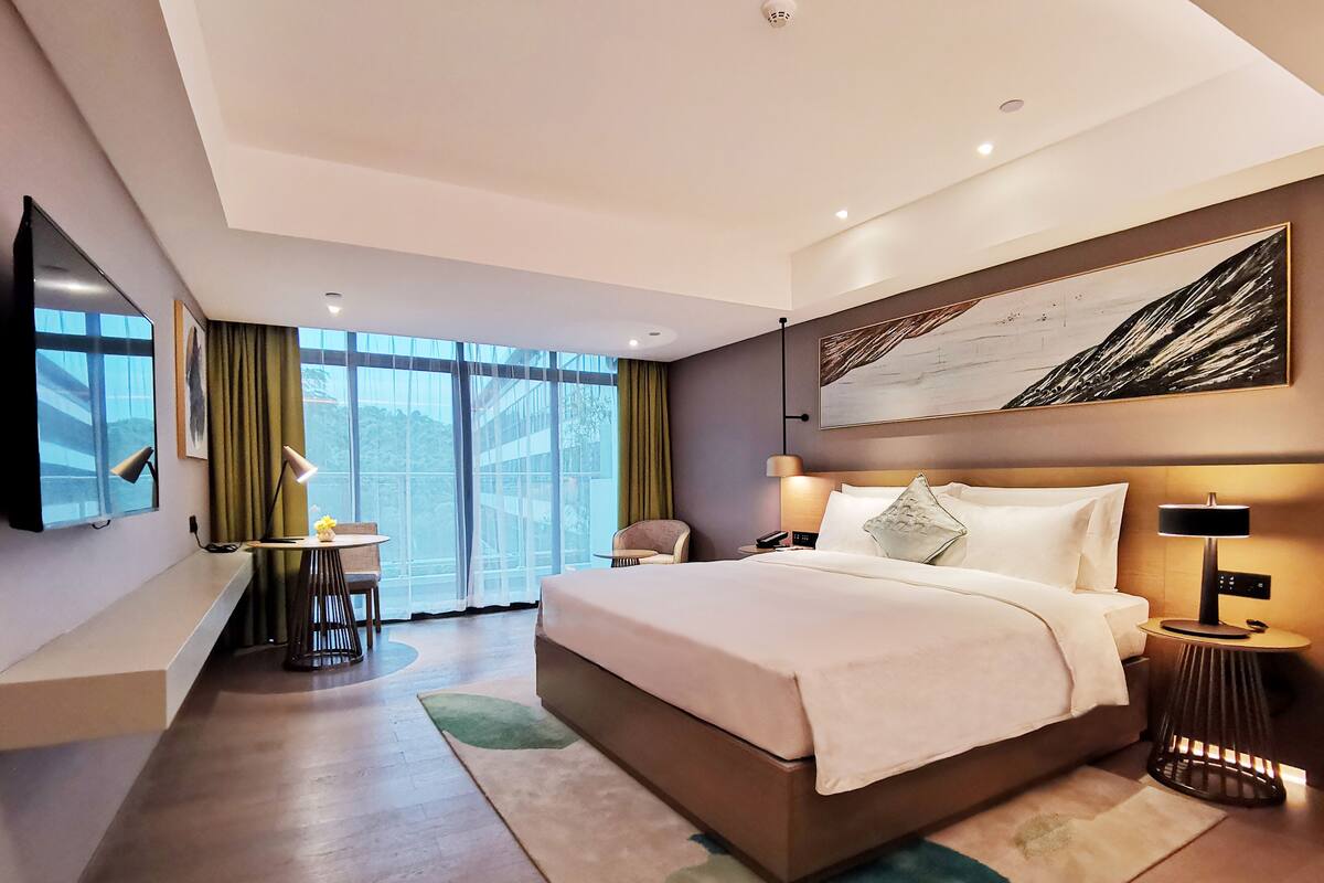 Ein modernes Hotelzimmer mit einem großen Bett, einem Schreibtisch mit Stuhl, einem Fernseher und einem großen Fenster mit Vorhängen.