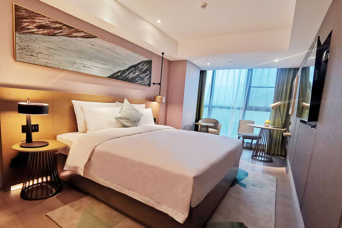 Ein modernes Hotelzimmer mit einem großen Bett, einem Nachttisch mit Lampe, einem kleinen Tisch mit Pflanze und Blick ins Freie durch große Fenster.