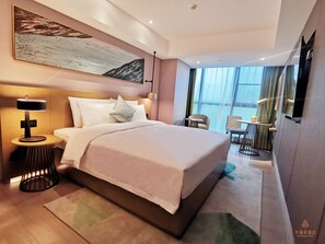 Habitación de hotel moderna con una cama grande, una mesita de noche con lámpara, una pequeña mesa con una planta y vistas al exterior a través de grandes ventanales.