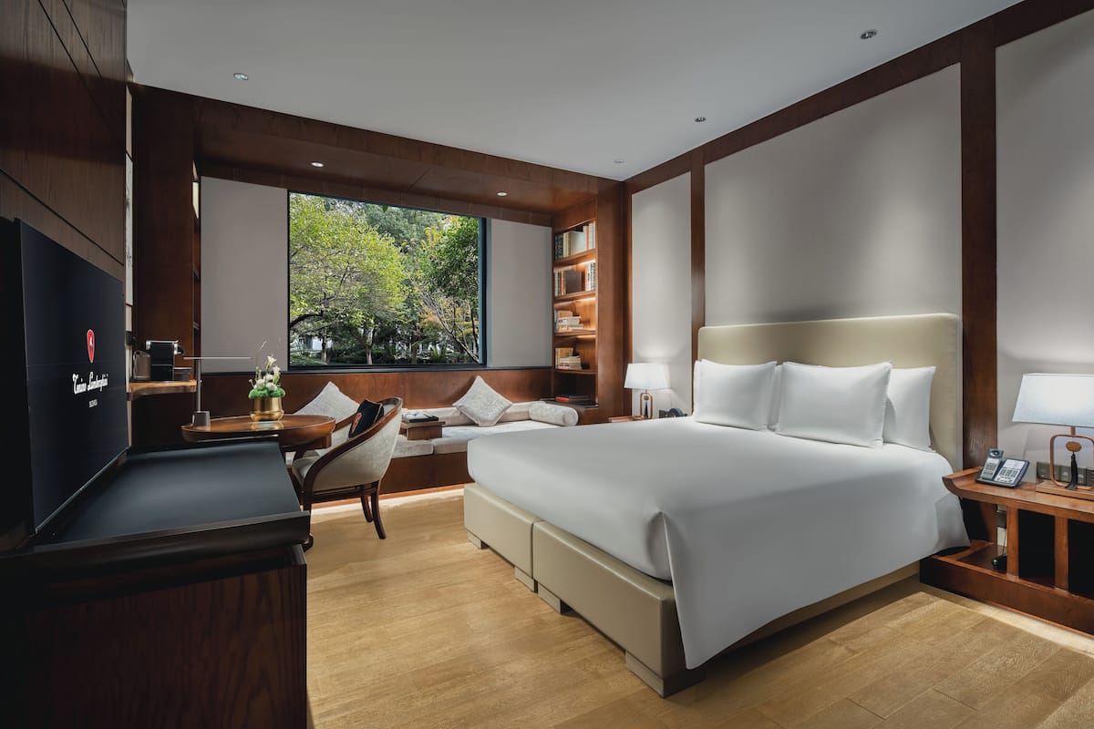 Une chambre d’hôtel moderne, dotée d’un grand lit, d’un bureau, d’une chaise, d’une bibliothèque et d’une grande fenêtre donnant sur la verdure.