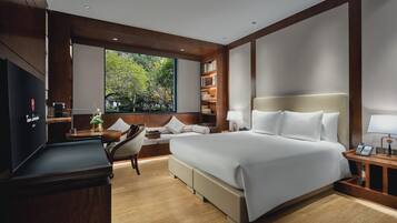 Quarto de hotel moderno com uma cama grande, uma escrivaninha, uma cadeira, uma estante de livros e uma janela ampla com vista para árvores.