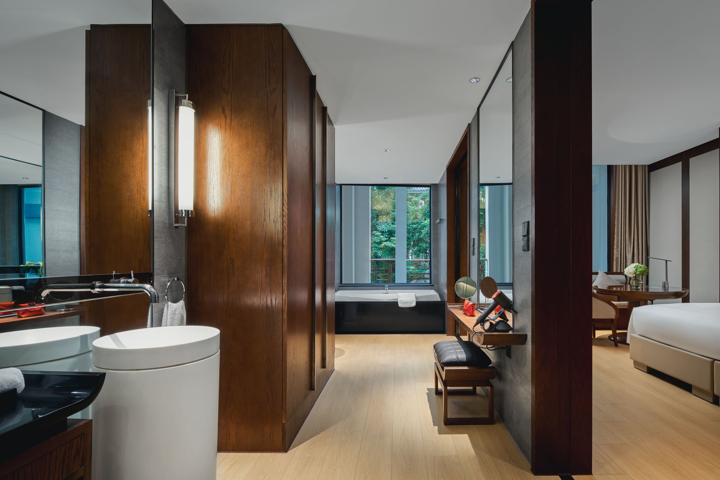 A modern hotel room with a large bed, a desk with a chair, a bathroom with a sink and mirror, and a large window with a view of trees.