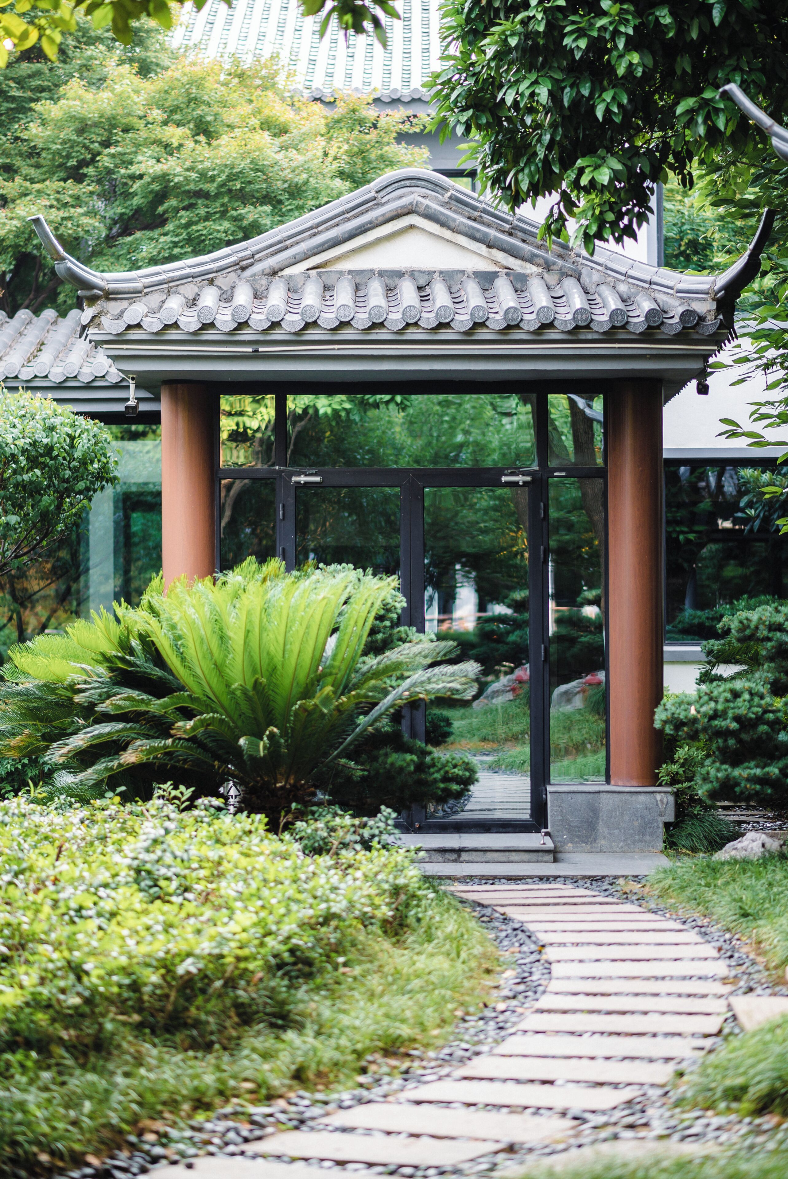 Un pavillon de style japonais traditionnel, doté d’un toit en tuiles, entouré d’une végétation luxuriante et bordé d’un sentier en pierre.