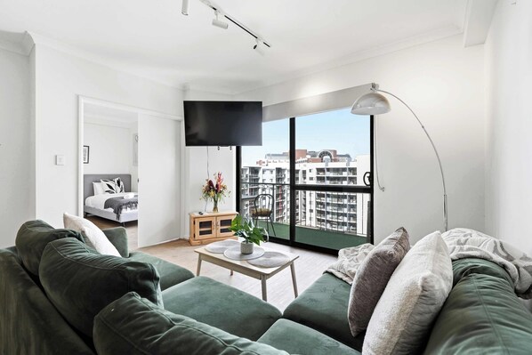 A modern living room with a green sofa, a small table with a vase, and a TV on the wall.