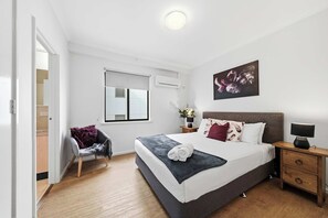 Un dormitorio con cama, una silla, una mesita de noche, una lámpara, un cuadro y una ventana con persianas.