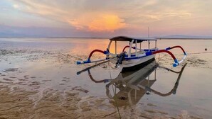 A small boat with a canopy and blue and red paddles on a calm body of water during sunset. - Akaya Bali (Denpasar)