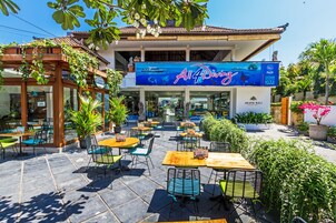 Un área de café al aire libre con mesas y sillas, un letrero grande que dice 'All 4 Diving', y un edificio con techo de tejas y plantas.