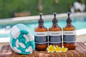 Three bottles of Sensatia Botanicals products, a rolled towel, and two flowers on a wicker surface by a pool.