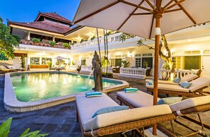 A pool area with lounge chairs and umbrellas, a statue, and a building with balconies and plants. - Akaya Bali (Denpasar)
