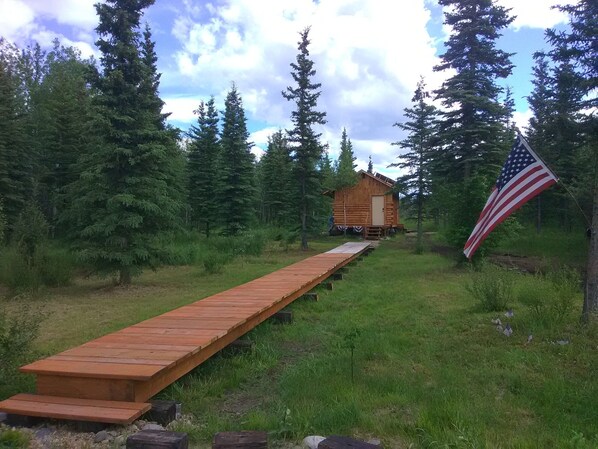 Une cabane en bois avec une terrasse en bois qui y mène, entourée d’une forêt dense et sur laquelle flotte un drapeau américain.