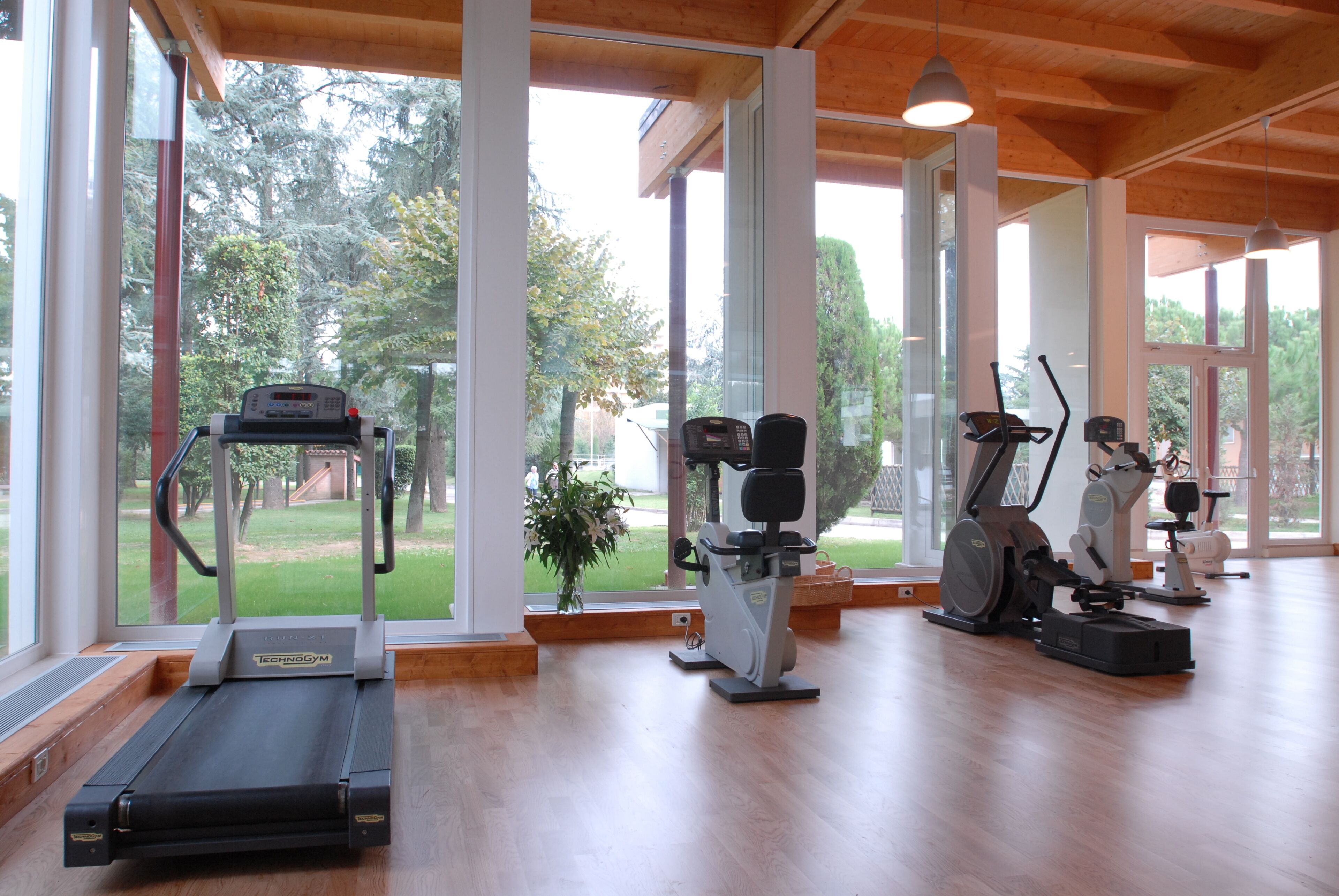 A gym with a treadmill, elliptical, and stationary bike, large windows, and wooden flooring.
