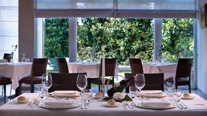 Une table à manger dressée pour un repas, avec une bouteille de vin, des verres et des assiettes, et une vue sur la verdure à travers de grandes fenêtres.