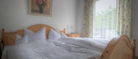 Cama doble con ropa blanca, cabecera de madera, mesita de noche con lámpara y ventana con cortinas florales.