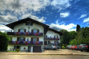 Un bâtiment à deux étages, doté de balcons ornés de fleurs, une enseigne indiquant « Gästehaus Meren », et des voitures garées au premier plan.