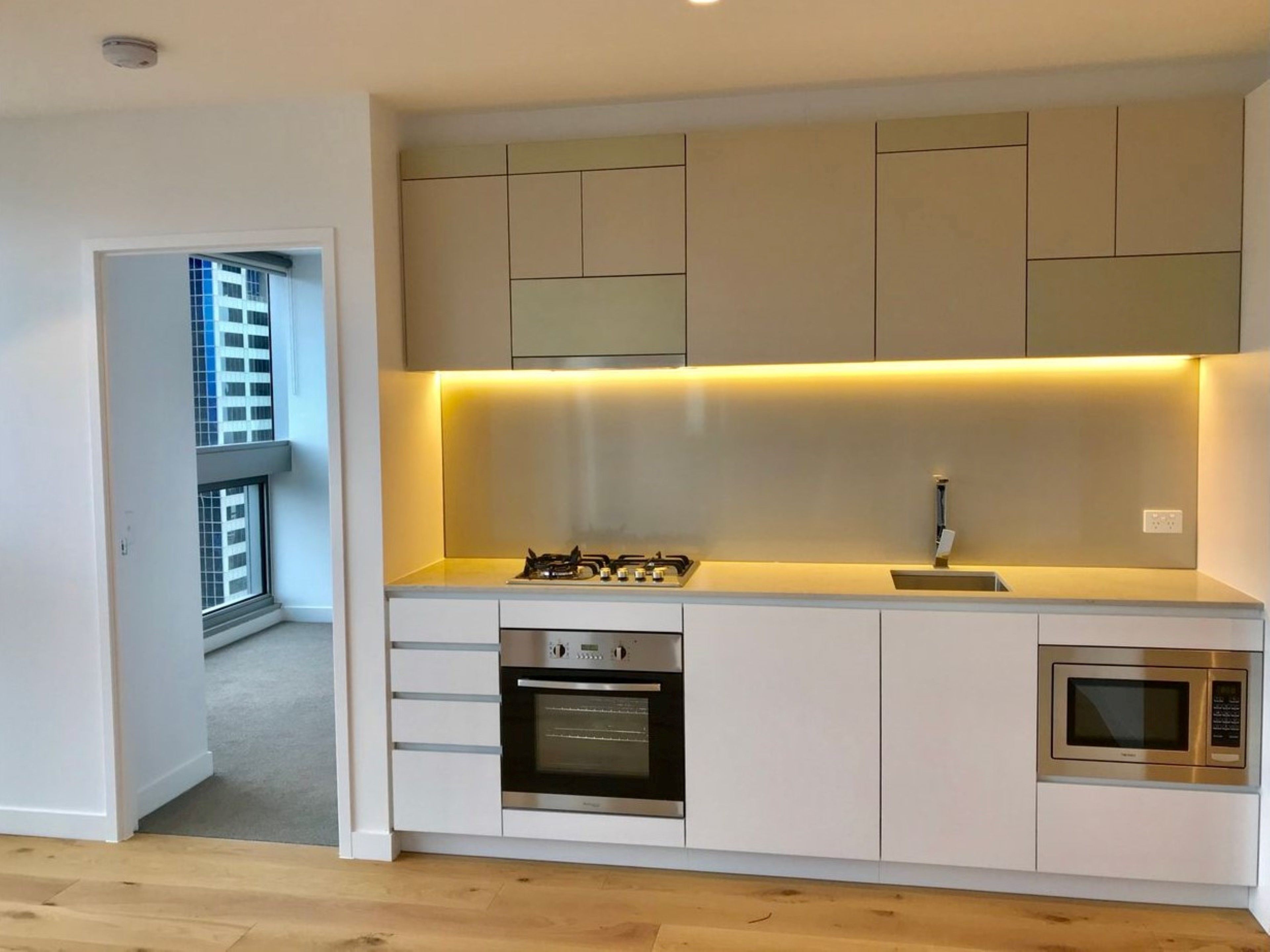 Une cuisine moderne équipée d’une cuisinière à gaz, d’un four et d’un micro-ondes, avec des meubles blancs et une vue sur un bâtiment par une porte ouverte.