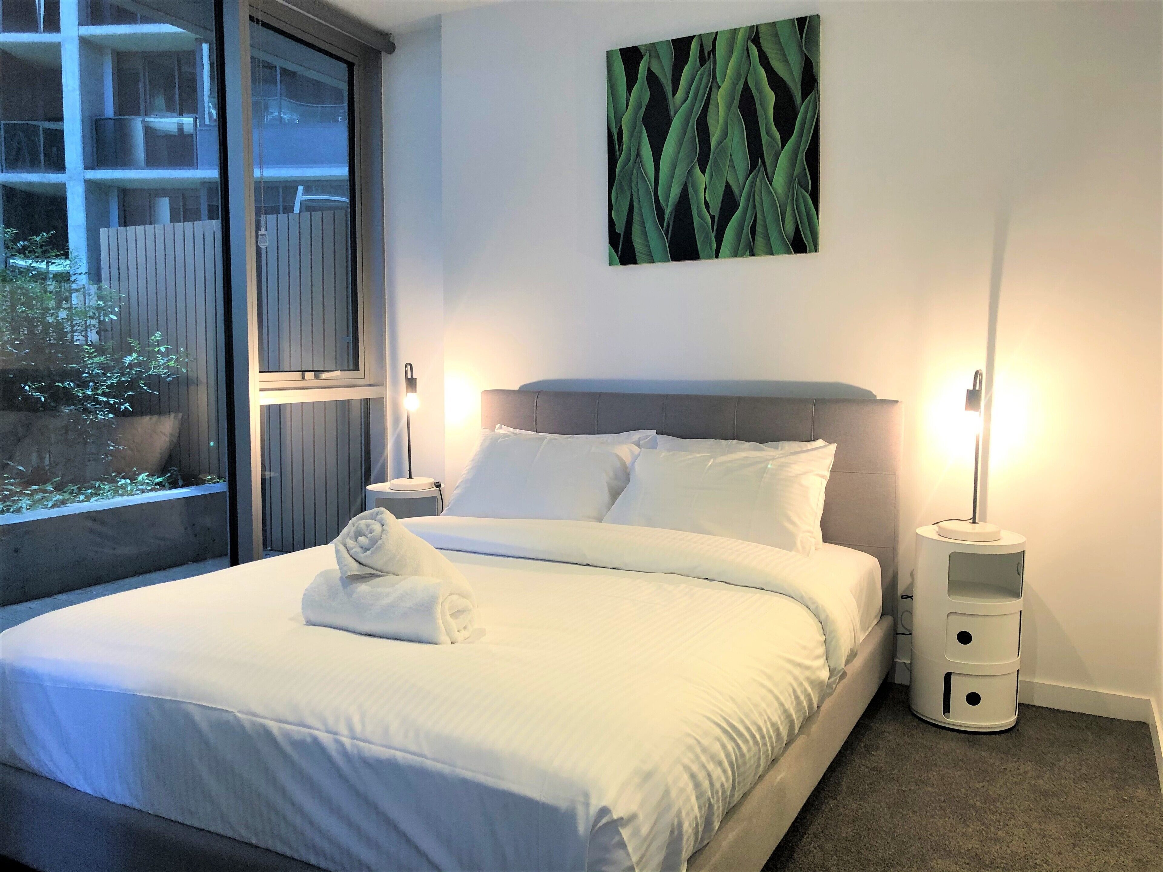A neatly made bed with white linens and a rolled towel, a bedside lamp, and a nightstand with a lamp.