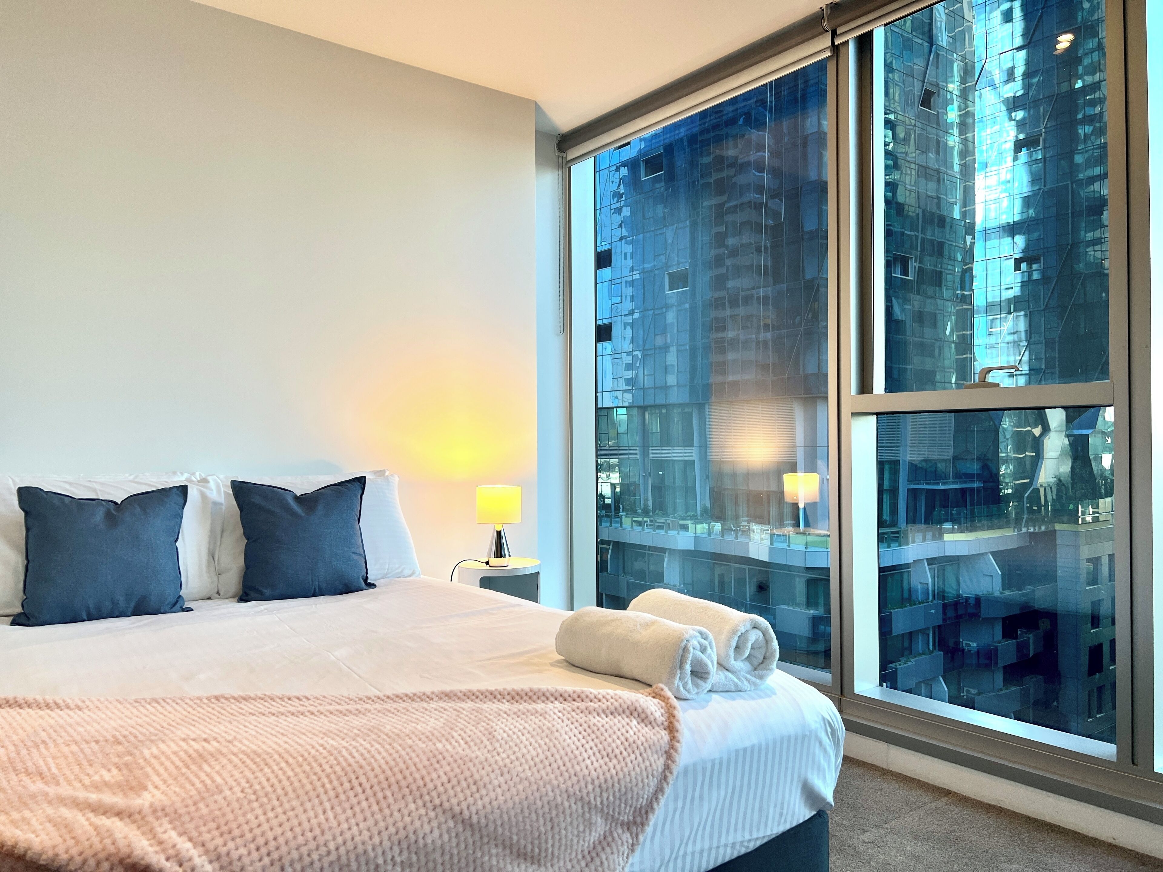 Une chambre d’hôtel avec un grand lit, deux oreillers, une lampe de chevet, une serviette roulée et une grande fenêtre donnant sur la ville.