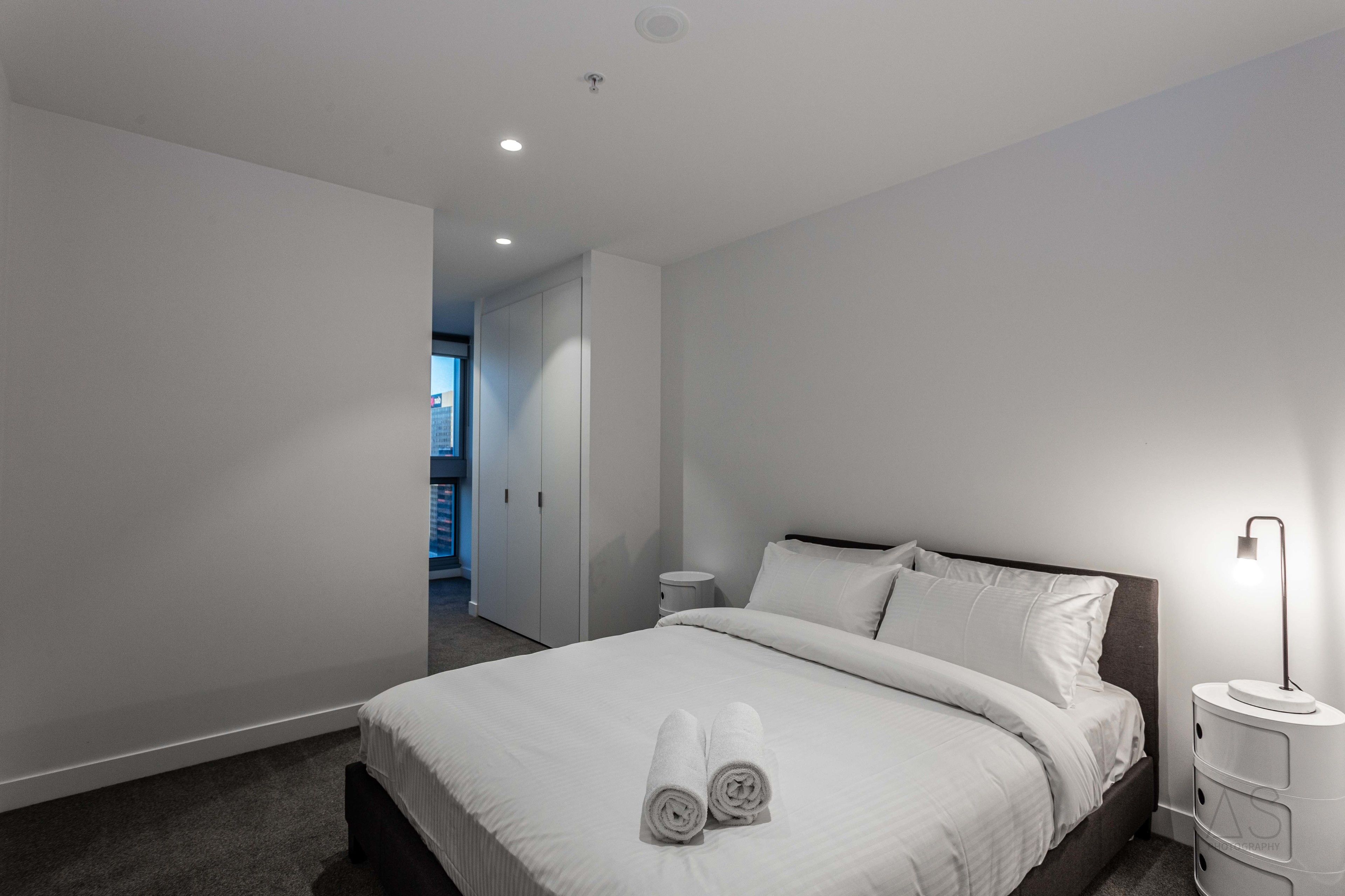 A modern hotel room with a large bed, white linens, a bedside table, and a nightstand with a lamp.