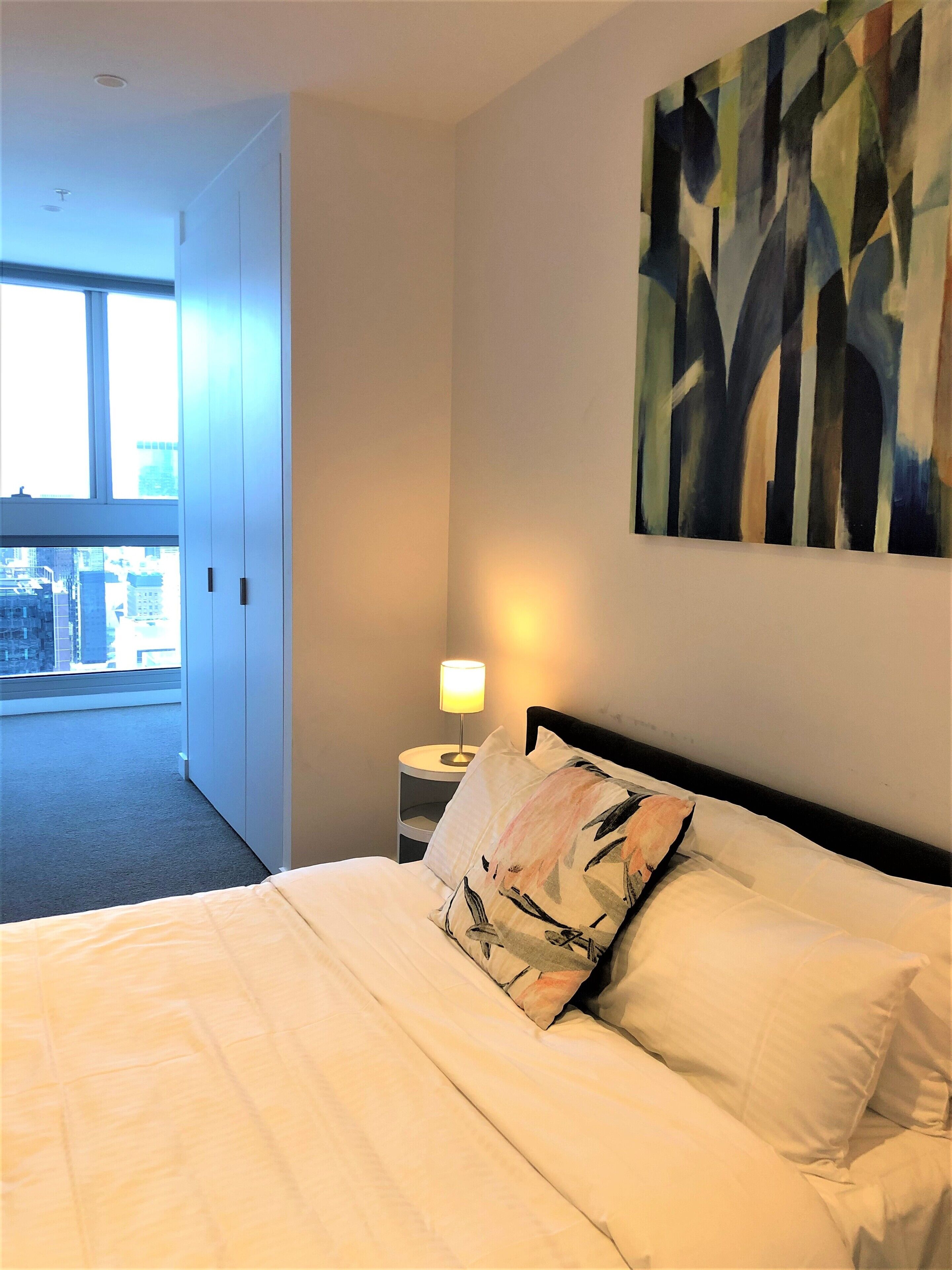 Una habitación de hotel con cama, mesita de noche con lámpara, un cuadro en la pared y vistas de la ciudad a través de un ventanal.
