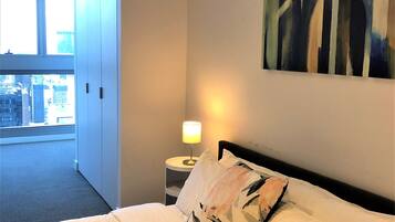 Una habitación de hotel con cama, mesita de noche con lámpara, un cuadro en la pared y vistas de la ciudad a través de un ventanal.