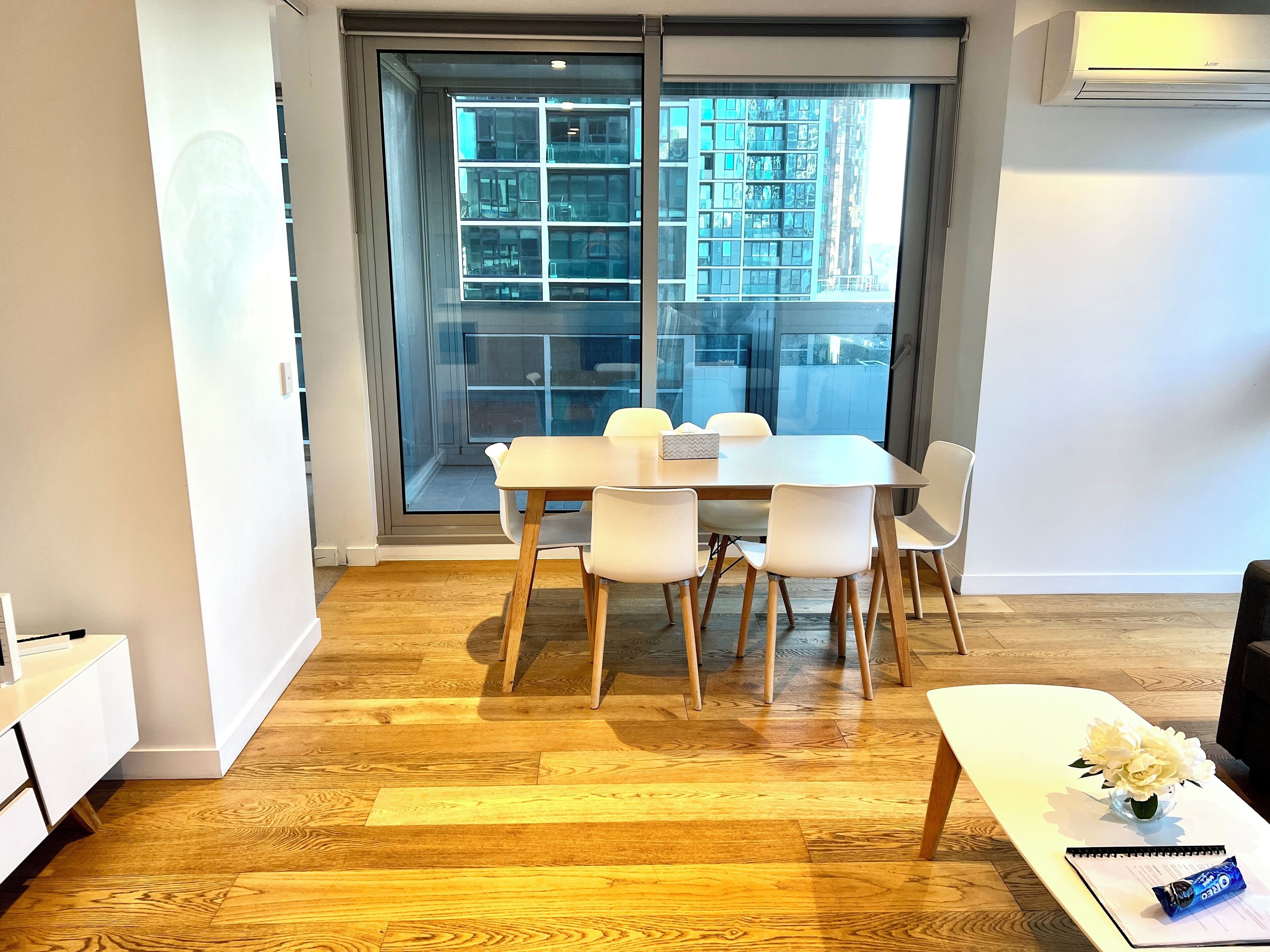 A modern dining area with a wooden table and white chairs, a white console table with a vase and a notebook, and a large window with a view of a cityscape.