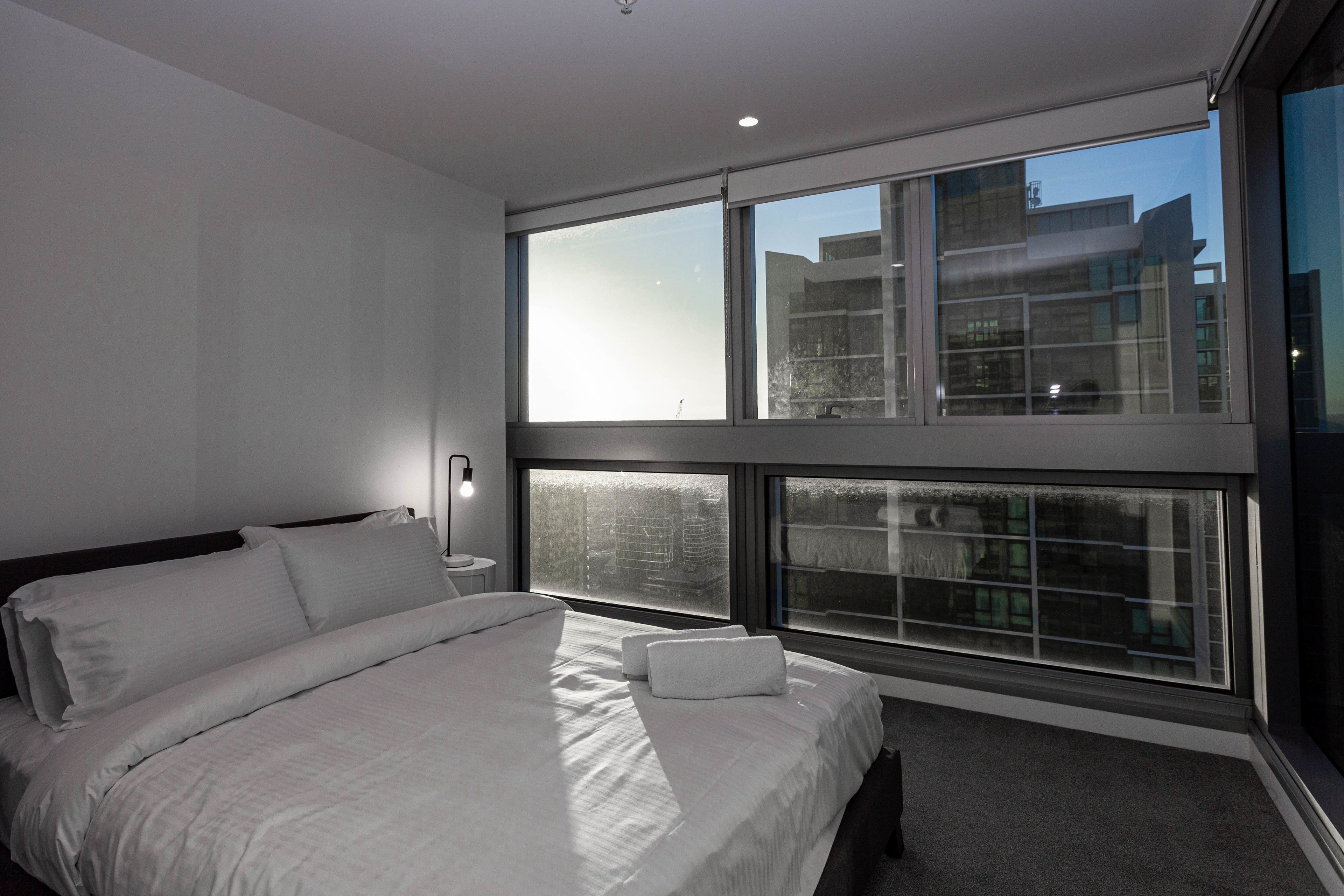 A modern hotel room with a large bed, a bedside table, and a view of the city through large windows.