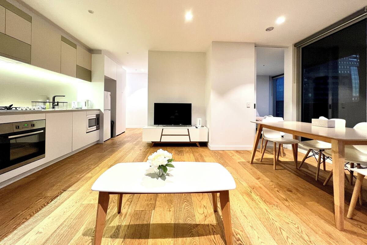 A modern kitchen with built-in appliances, a dining area with a wooden table and chairs, and a TV mounted on the wall.