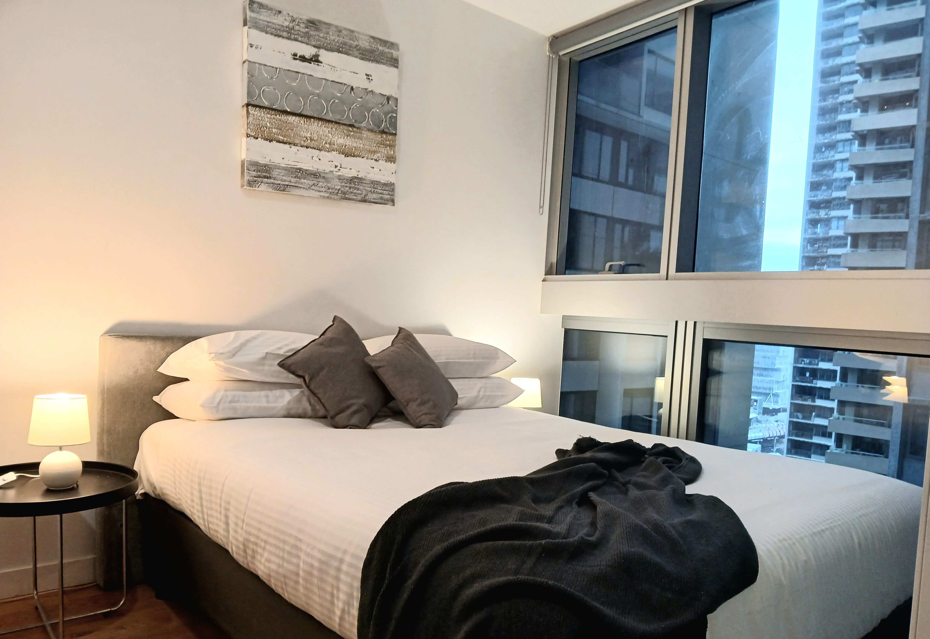 A hotel room with a bed, a nightstand with a lamp, a window with a city view, and a wall art piece.