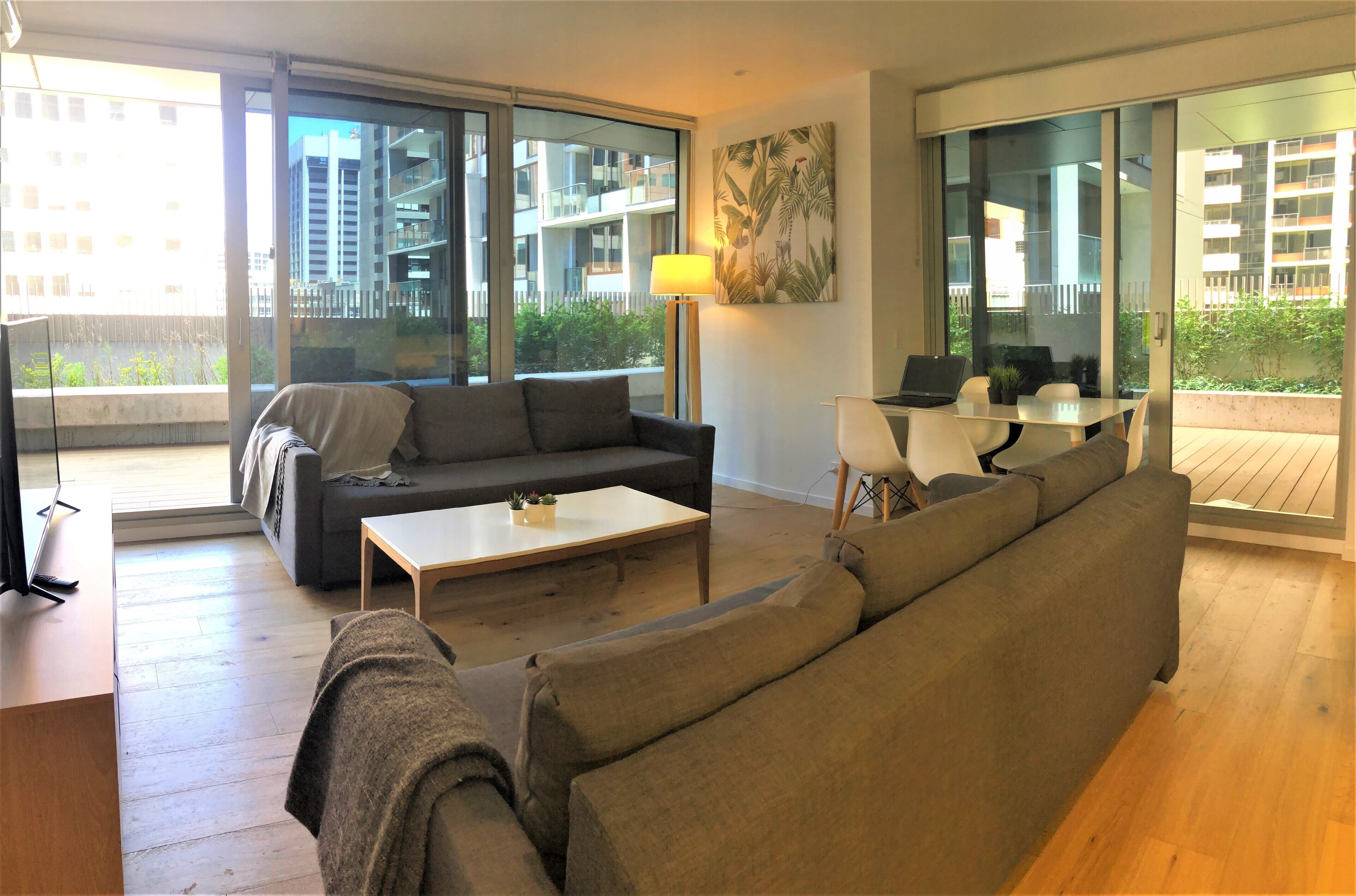 A modern living room with a sofa, a coffee table, and a desk with a computer. Large windows offer views of a cityscape and greenery.