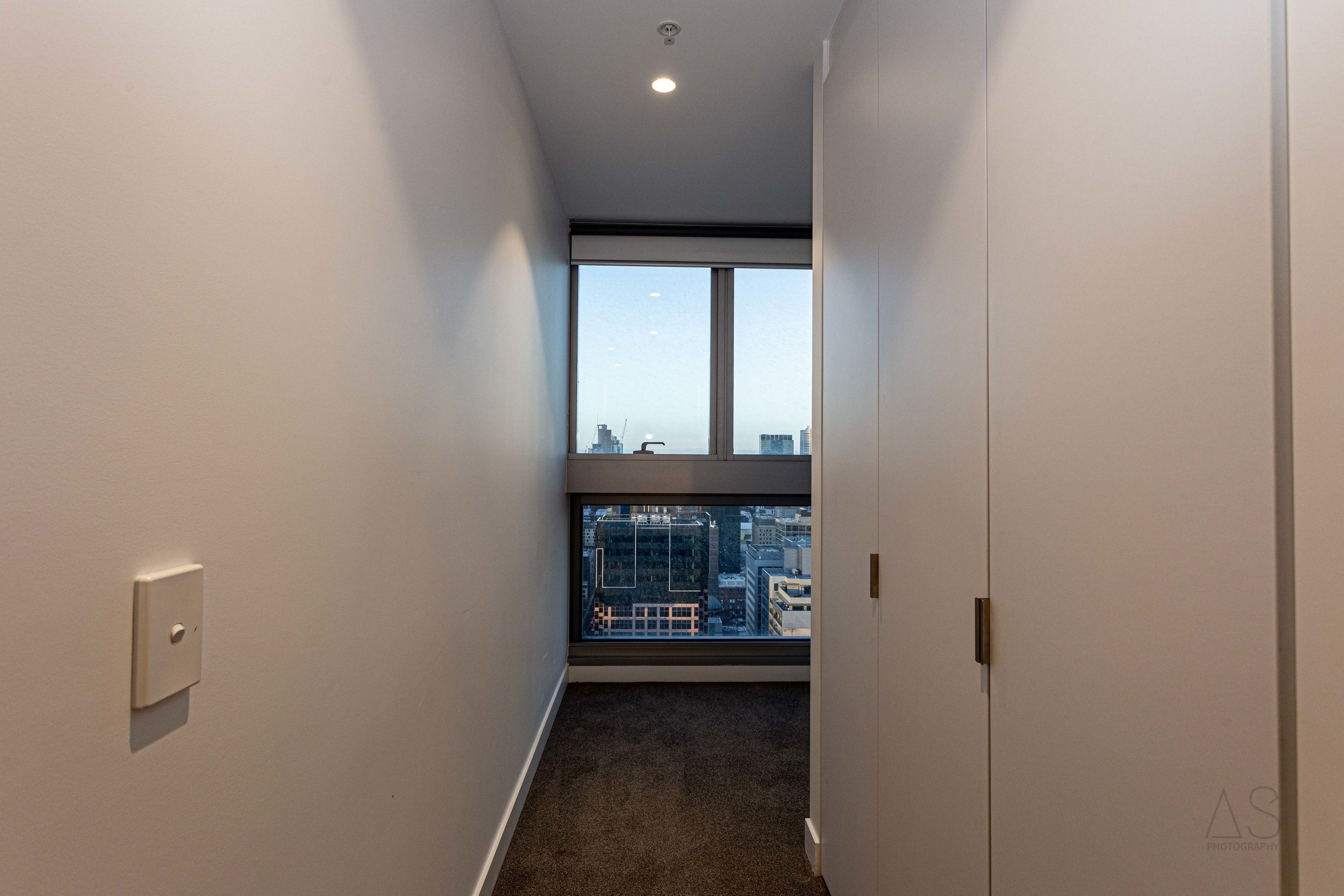 A narrow hallway with a carpeted floor, white walls, and a large window at the end.