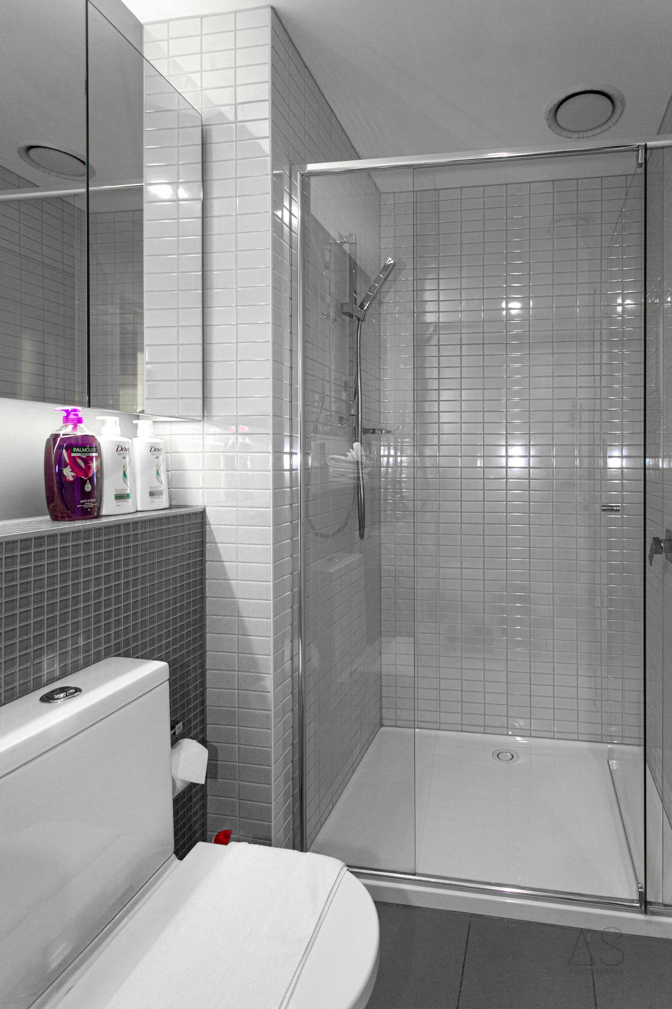 A modern bathroom with a glass-enclosed shower, white tiles, a toilet, and a sink with toiletries.