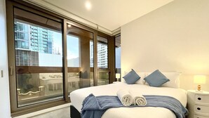 Une chambre d’hôtel moderne dotée d’un grand lit, offrant une vue imprenable grâce à de larges fenêtres du sol au plafond, et comprenant une table de chevet avec une lampe.