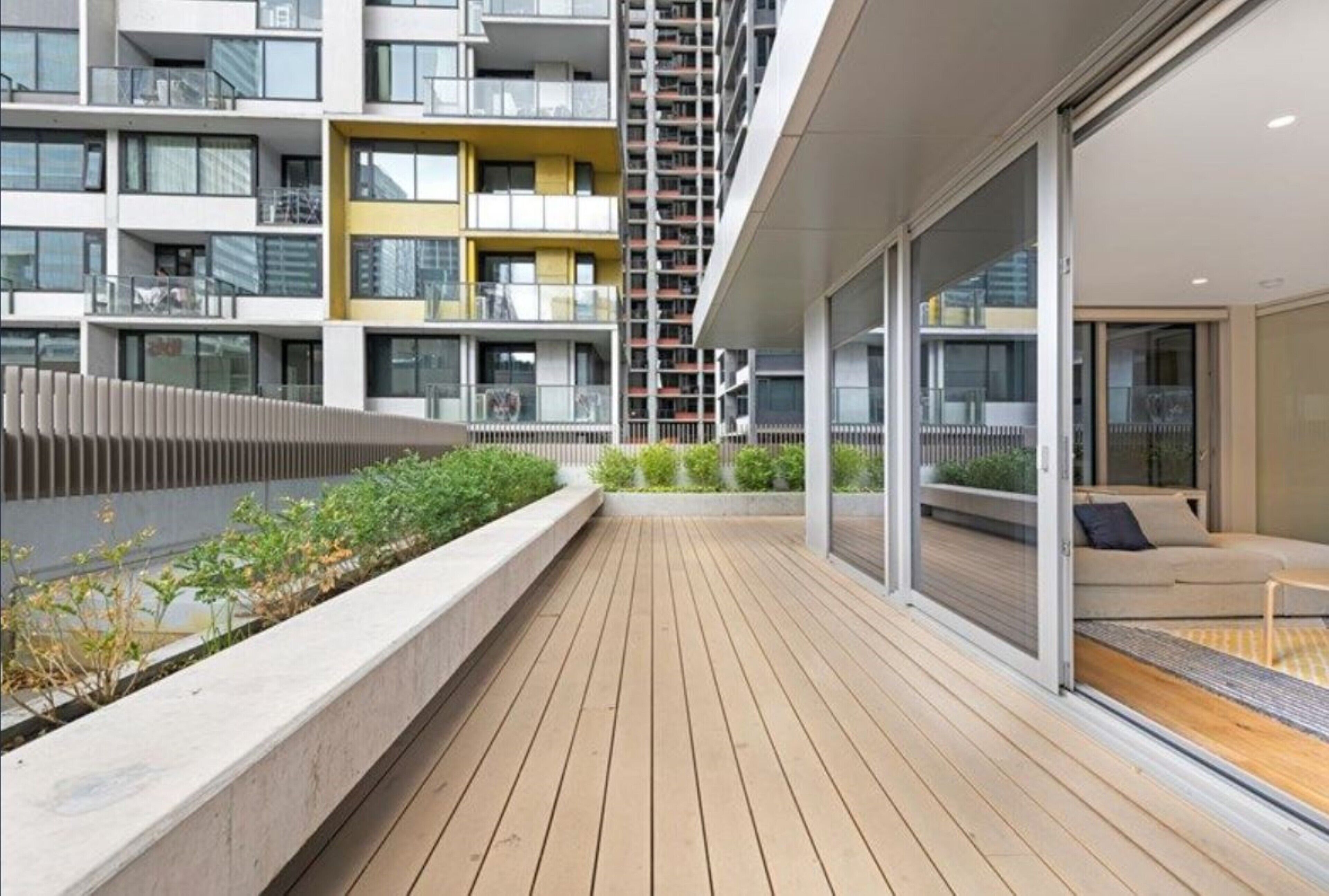 Un edificio de apartamentos moderno con balcones, una terraza de madera y una puerta de cristal que da a un área de estar.
