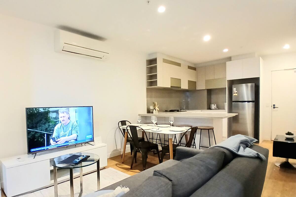 A modern living room with a grey sofa, a small coffee table, and a flat-screen TV. There is a dining area with a white table and chairs, and a kitchen with white cabinets and a stainless steel refrigerator.