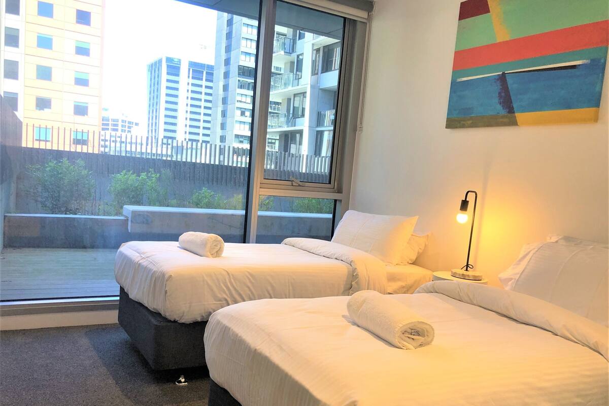 A hotel room with two beds, a large window, a painting, and a nightstand with a lamp.