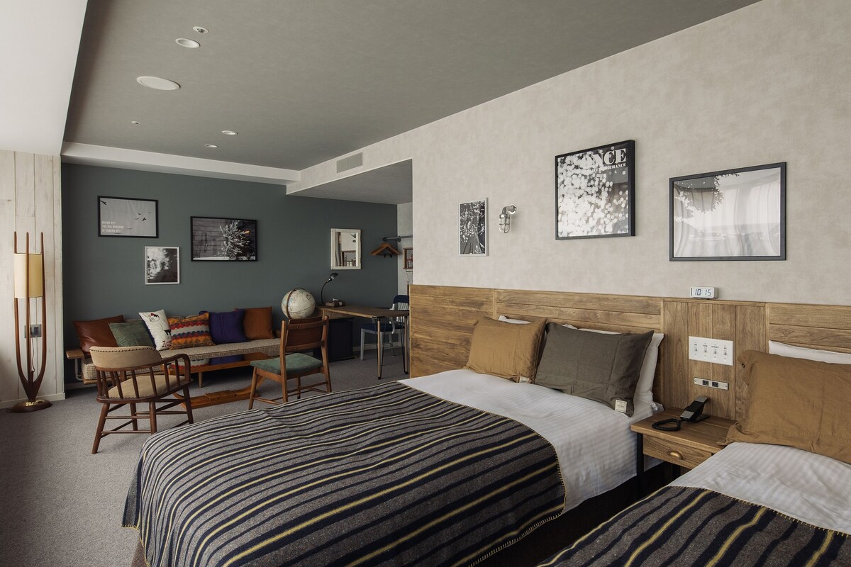 Habitación de hotel con dos camas, un escritorio, una silla y un sofá. Incluye luces empotradas en la pared y obras de arte.
